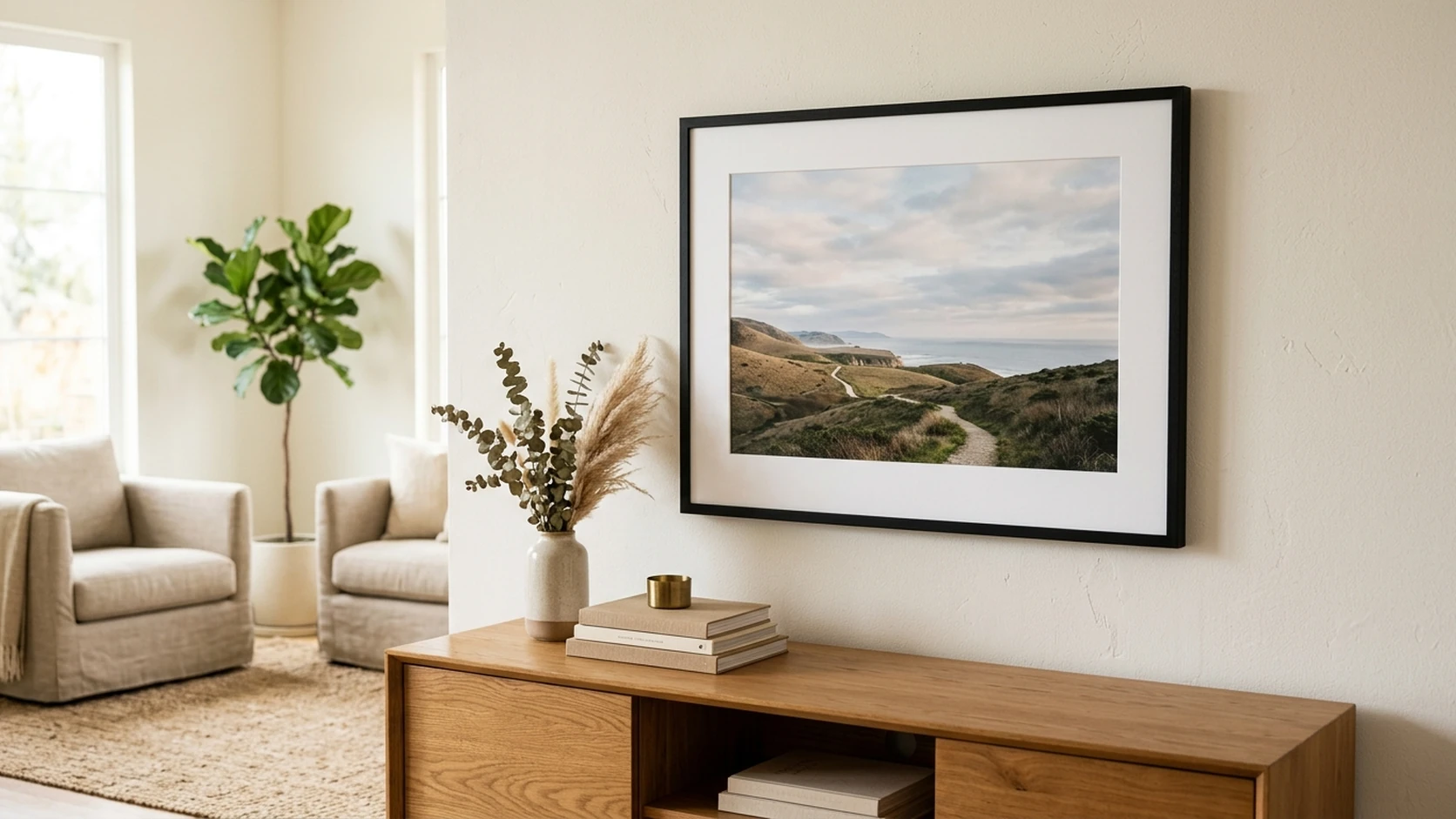 A black-framed, matted coastal landscape print hanging above an oak sideboard in a bright living room with cream armchairs