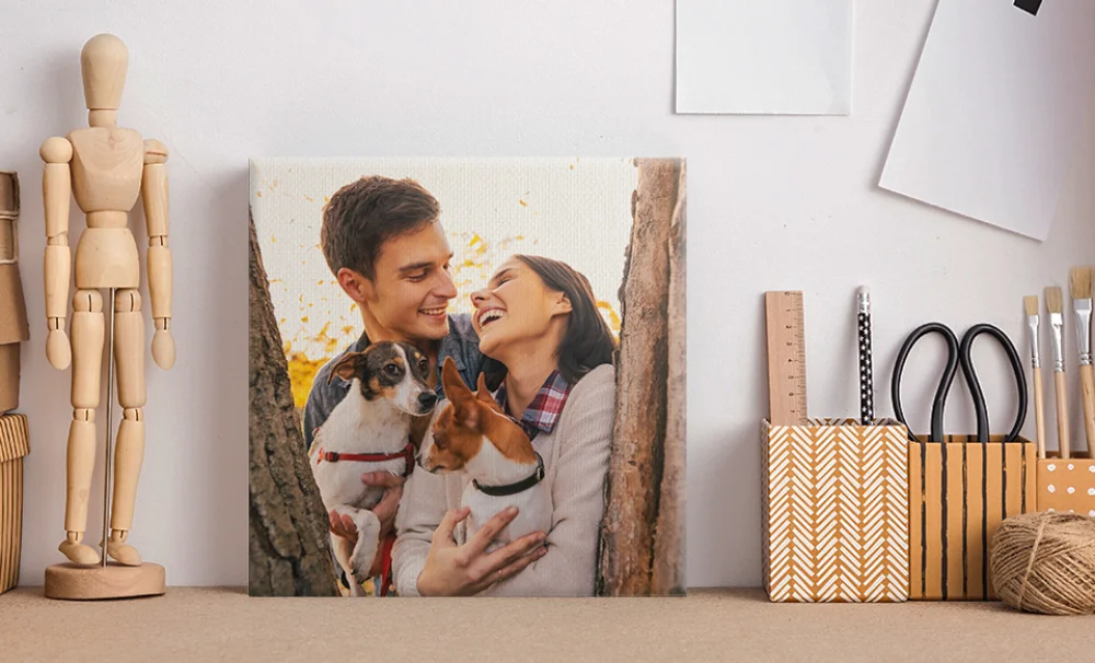 Canvas print of a smiling couple holding two small dogs, propped on a desk with art supplies